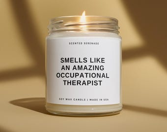 May include: A clear glass jar candle with a white label featuring the text Scented Serenade, Smells Like An Amazing Occupational Therapist, and Soy Wax Candle Made In USA. The candle has a lit wick and contains off-white wax.