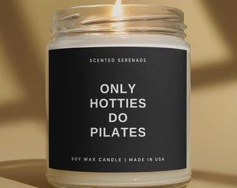 May include: A clear glass jar candle with a lit wick and a black label. The label features the white text SCENTED SERENADE, ONLY HOTTIES DO PILATES, and SOY WAX CANDLE | MADE IN USA. The candle contains off-white wax and is filled to the top.