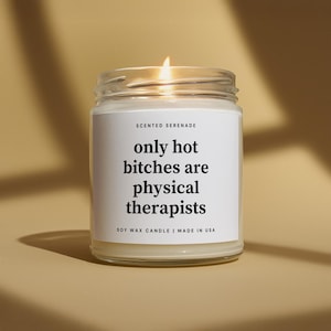May include: A clear glass jar soy wax candle with a white label featuring the black text Scented Serenade, only hot bitches are physical therapists, and Soy Wax Candle | Made in USA. The candle has a lit wick and a visible flame.