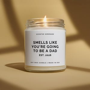 May include: A lit soy wax candle in a glass jar. The candle has a white label with the text "SMELLS LIKE YOU'RE GOING TO BE A DAD EST. 2026". The candle is made in the USA.