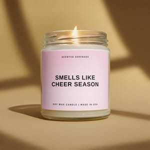 May include: A clear glass jar candle with a lit wick and a light pink label. The label features the text SCENTED SERENADE, SMELLS LIKE CHEER SEASON, and SOY WAX CANDLE | MADE IN USA in black sans-serif font. The candle wax is off-white.