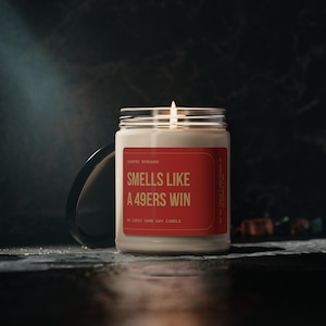 May include: A clear glass jar candle with a white wax fill and a lit wick. The label is red with yellow text that reads: SCENTED SERENADE, SMELLS LIKE A 49ERS WIN, MY LUCKY GAME DAY CANDLE. The candle has a black lid resting beside it.
