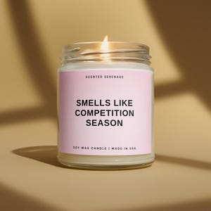 May include: A lit soy wax candle in a clear glass jar with a light pink label. The label features the text Scented Serenade, Smells Like Competition Season, and Soy Wax Candle | Made in USA printed in black sans-serif font.
