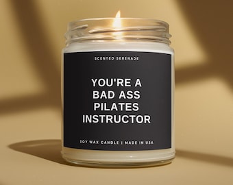 May include: A clear glass jar soy wax candle with a lit wick. The black label features the white text: SCENTED SERENADE, YOU'RE A BAD ASS PILATES INSTRUCTOR, and SOY WAX CANDLE | MADE IN USA. The candle emits a warm glow from the flame.