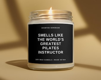 May include: A clear glass jar candle with a lit wick and a black label. The label features white text reading: SCENTED SERENADE, SMELLS LIKE THE WORLD'S GREATEST PILATES INSTRUCTOR, SOY WAX CANDLE | MADE IN USA. The candle contains white wax.