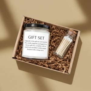 A gift set containing a candle and a match holder, placed in a cardboard box filled with shredded paper.