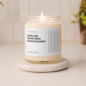 Peut inclure: Une bougie en cire de soja allumée dans un pot en verre transparent avec une étiquette blanche. L'étiquette indique : Scented Serenade, Smells Like World's Best Dental Assistant, Hand-poured Soy Wax Candle, 50-60 Hours Burning Time, Eco-Friendly & Non-Toxic, KEEP THAT SHIT UP.