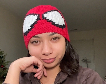 Puede incluir: Una persona lleva una gorra roja de punto con un diseño de Spiderman blanco y una sudadera con capucha marrón con cremallera con el texto "REEF DK" en la parte delantera.