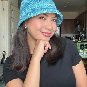 Peut inclure: Un chapeau seau en crochet bleu clair avec un motif de point texturé. Le chapeau a un bord légèrement incliné vers le bas. Le chapeau est porté par une personne portant un t-shirt noir.