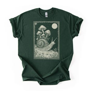 Può includere: T-shirt verde scuro con un'illustrazione dettagliata di una lumaca che trasporta funghi sotto la luna piena. Il disegno è incorniciato da un bordo decorativo. La maglietta è realizzata in un materiale morbido.