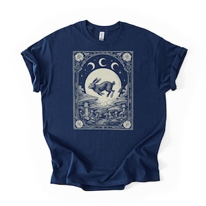 Puede incluir: Camiseta azul marino con un gráfico detallado de un conejo saltando frente a una luna llena, rodeado de setas y elementos celestiales. El diseño está enmarcado dentro de un borde ornamentado, creando una estética mística y caprichosa.