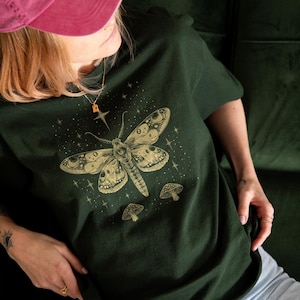 以下が含まれることがあります： モス、キノコ、星の精巧なイラストが描かれた濃い緑色のTシャツ。モスとキノコは薄茶色です。半袖Tシャツはカジュアルな服装です。