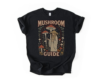 May include: Black t-shirt featuring a whimsical illustration. The design includes a figure with a staff, surrounded by mushrooms and foliage, with the words "MUSHROOM GUIDE" in a decorative font.