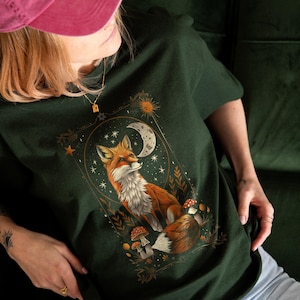 Könnte beinhalten: Dunkelgrünes T-Shirt mit einer detaillierten Illustration eines Fuchses mit weiß-orangefarbenem Fell, vor dem Hintergrund eines Halbmonds, Sternen und Pilzen. Das Design ist von einem goldenen Rahmen umgeben und schafft eine skurrile, von der Natur inspirierte Ästhetik.