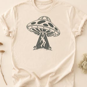 May include: A cream-colored short-sleeve t-shirt featuring a dark gray graphic print. The design depicts a flying saucer hovering above the ground, beaming light down onto a person who is floating upward, flanked by two small mushrooms on the ground.