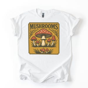 Puede incluir: Camiseta blanca con un diseño rectangular marrón con setas. La palabra "MUSHROOMS" está en texto marrón encima de las setas. Las setas son rojas y blancas con tallos marrones.