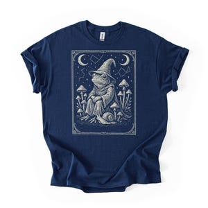 May include: Navy blue t-shirt featuring a detailed illustration of a frog wizard wearing a hat and robe, surrounded by mushrooms and a snail. The design includes celestial elements like stars and crescent moons, all within a decorative frame.