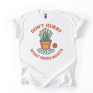 Op de afbeelding: Wit T-shirt met een grafisch ontwerp met een plant in een pot en de tekst "DON'T HURRY WHAT NEEDS ROOTS" in het oranje. Het ontwerp bevat een kleine klok en wortels.