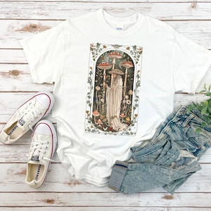 May include: White t-shirt featuring a whimsical illustration of a figure among mushrooms. The design includes a person in a long white robe and a hat, surrounded by tall mushrooms with red and white caps. The shirt is paired with white sneakers and blue jeans.