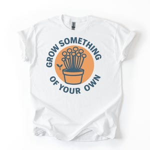 Puede incluir: Camiseta blanca con un diseño gráfico circular. El diseño presenta una maceta con plantas estilizadas y el texto "GROW SOMETHING OF YOUR OWN" en una combinación de colores azul y naranja. La camiseta está hecha de un material suave.