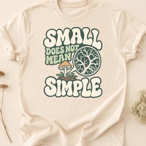 May include: A cream-colored t-shirt featuring a graphic with the text "SMALL DOES NOT MEAN SIMPLE" in dark green and light green. The design includes a small orange mushroom and a magnifying glass showing a detailed root system.