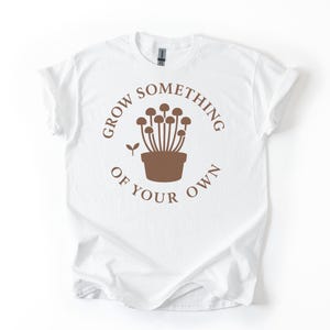 Op de afbeelding: Wit t-shirt met een bruine afbeelding van paddenstoelen in een pot en de tekst "GROW SOMETHING OF YOUR OWN" in een cirkel. Het t-shirt heeft opgerolde mouwen en is gemaakt van zacht materiaal.