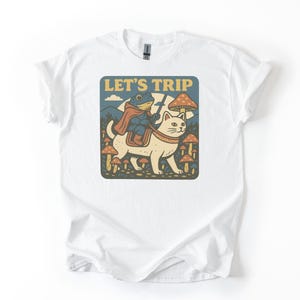 Pode incluir: Camiseta branca com um gráfico caprichoso. O design inclui um gato carregando um sapo, com o texto "LET'S TRIP" em uma fonte retrô. A imagem também apresenta cogumelos e um fundo de montanha.