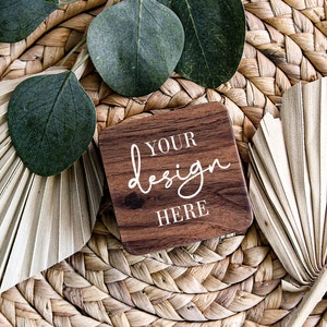 May include: A wooden coaster with the text "YOUR design HERE" in white script. The coaster is surrounded by dried palm leaves and green eucalyptus leaves on a woven wicker surface.