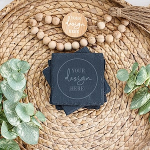Peut inclure: Quatre sous-verres en ardoise noire avec un motif rond blanc qui dit "YOUR design HERE" sur un plateau en paille tressée avec une guirlande de perles en bois et du feuillage vert.