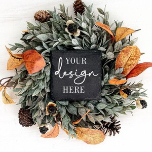Puede incluir: Una corona de otoño con un letrero de pizarra negra que dice "YOUR design HERE". La corona está hecha de hojas verdes, piñas marrones y hojas naranjas.