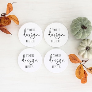 Puede incluir: Cuatro posavasos redondos blancos con el texto "Your design here" en escritura negra. Los posavasos están sobre una superficie de madera blanca con hojas de otoño y dos pequeñas calabazas blancas.