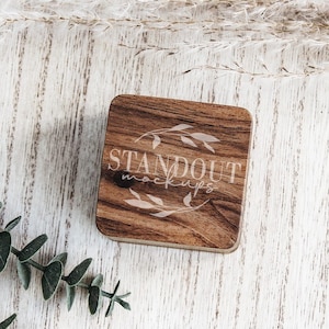 May include: A wooden square sign with the text "STANDOUT MOCKUPS" in white lettering with a floral design. The sign is on a white wooden surface with green eucalyptus branches.