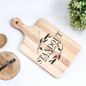 May include: A wooden cutting board with a handle, featuring the text "STANDOUT mockups" in a black script font with a floral design. The board is on a white surface.