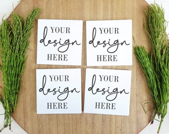 Puede incluir: Cuatro posavasos cuadrados blancos con el texto "Your design here" escrito en letra cursiva negra en cada posavasos. Los posavasos están dispuestos en una cuadrícula de 2x2 sobre una superficie de madera.