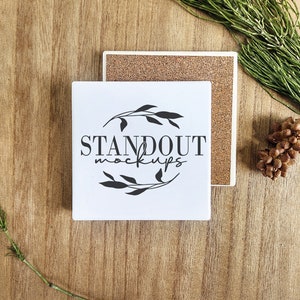 May include: White ceramic coaster with a black floral design and the text "STANDOUT MOCKUPS". The coaster is on a wooden surface with a brown cork coaster underneath.
