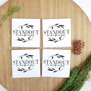 Op de afbeelding: Vier witte vierkante onderzetters met zwarte tekst en een eenvoudig bloemmotief. De tekst op elke onderzetter luidt "STANDOUT MOCKUPS". De onderzetters zijn in een vierkant op een houten oppervlak gerangschikt.