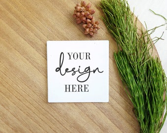 Puede incluir: Una tarjeta blanca cuadrada con el texto "YOUR design HERE" sobre una superficie de madera con ramas verdes y flores decorativas marrones.