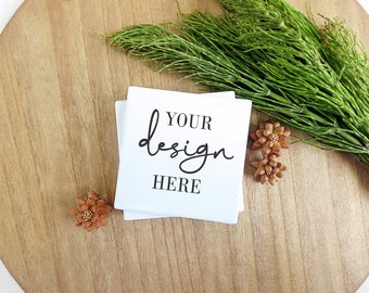 Puede incluir: Una pila de tarjetas cuadradas blancas con el texto "YOUR design HERE" en escritura negra. Las tarjetas están sobre una superficie de madera, con follaje verde y pequeños elementos decorativos marrones. La estética general es limpia y minimalista.