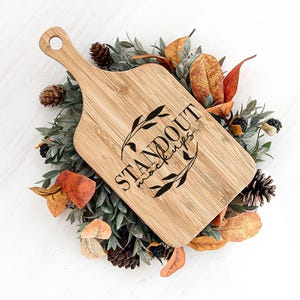 May include: A wooden cutting board with a handle, surrounded by fall foliage. The board is engraved with the text "Standout Mockups".