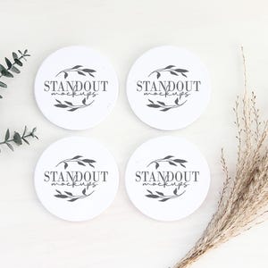 May include: Four white round coasters with the text "Standout Mockups" in black lettering and a simple floral design. The coasters are arranged in a square shape on a white surface with a sprig of eucalyptus and a bunch of dried pampas grass.