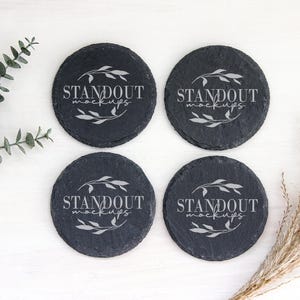 May include: Four round, dark gray slate coasters with the text "STANDOUT mockups" in a white, stylized font. Each coaster features a decorative leaf design. The coasters are arranged on a white surface with sprigs of greenery.
