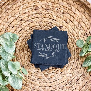 May include: Four black slate coasters with rounded edges, stacked on top of each other. The top coaster has the text "STANDOUT mockups" in a light gray color with a simple leaf design.