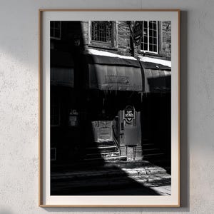 May include: Black and white photograph of a building with a cafe. The cafe has a sign that says "Cafe de Paris". The building has a dark awning and several windows. The image is framed in a wooden frame.