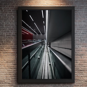 May include: Framed print of a modern architectural interior. The image features a long, reflective surface with a glass barrier, leading into a tunnel-like space. Overhead, linear lights illuminate the scene, with a red accent wall visible.