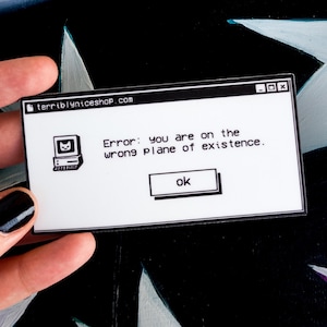 May include: A white sticker with a black border featuring a retro computer graphic and the text "Error: you are on the wrong plane of existence." The sticker also includes the text "terriblyniceshop.com" and an "ok" button.