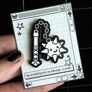 May include: A black and white enamel pin depicting a mace and a smiling sun-like face. The pin is on a white card with a grid pattern and the text "terriblyniceshop.com".