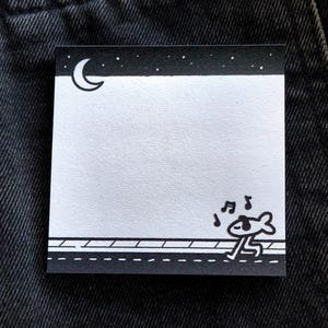 May include: Square sticky note with a black and white illustration. The top has a crescent moon and stars. The bottom features a road with a running fish character and musical notes. The background is a dark denim.