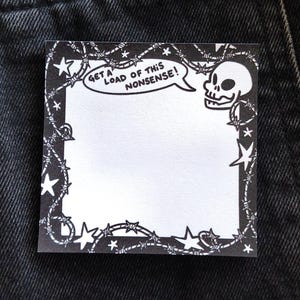 Könnte beinhalten: Quadratischer schwarz-weißer Notizblock mit einem Schädel- und Stacheldrahtrand. Der Notizblock hat den Satz "GET A LOAD OF THIS NONSENSE!" in einer Sprechblase. Der Notizblock liegt auf einer dunklen Jeansoberfläche.