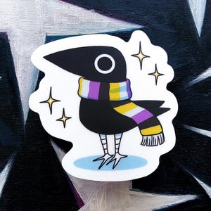 以下が含まれることがあります： ノンバイナリープライドのスカーフを身に着けた黒いカラスのステッカー。カラスは白い目を持ち、水色の楕円の上に立っています。4つの黄色の星がカラスを囲んでいます。ステッカーは白と黒の背景にあります。