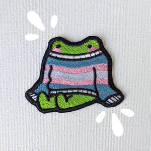 May include: Green frog wearing a blue, pink, and white striped sweater. The frog is sitting on a white background with white squiggles around it.